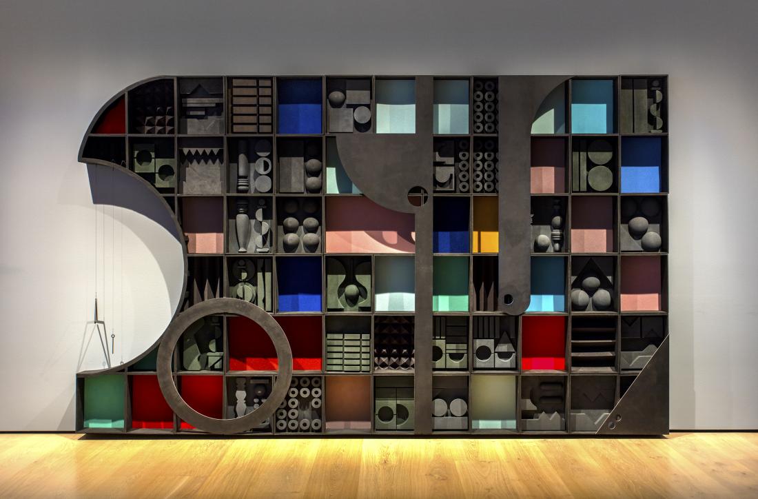 A large sculpture made of many rectangular cubbies filled with shaped acoustic foam stands flat against a wall. The depth of the cubbies casts shadows over flat red, blue, yellow, aqua, and pink foam panels. Flat, black pieces of wood in various shapes cover parts of the sculpture. The left side has a half circle cut out with a musical triangle hanging inside.