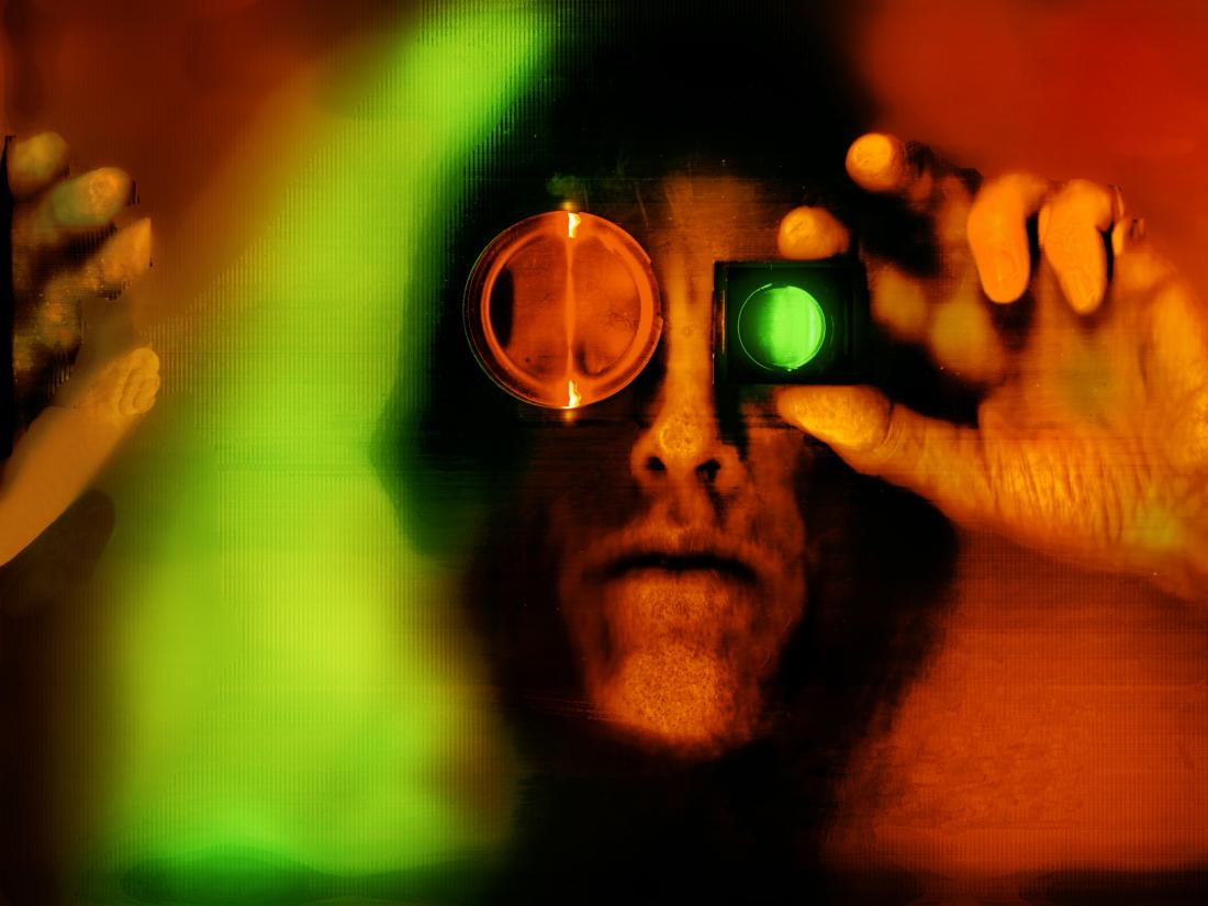A color self-portrait scanograph representing disintegration of  the body and vision.  The primary colors are green, red and black. My face is looking straight ahead and I am holding a monocular in front of my eye. The sides of my face are silhouetted in black. The image is very distorted and phased. This image is from my “Transmog Project” body of work. 