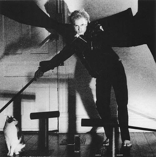 A portrait of a young man with blond hair and moustache wearing a leather motorcycle jacket and black pants. He is looking directly into the camera and leaning on a black metal staff which is positioned diagonally in the frame. There is a grey and white cat on the floor sitting next to his staff and is looking up at the young man. There are various objects positioned on the floor and the lighting is casting shadows on the wall behind the young man.