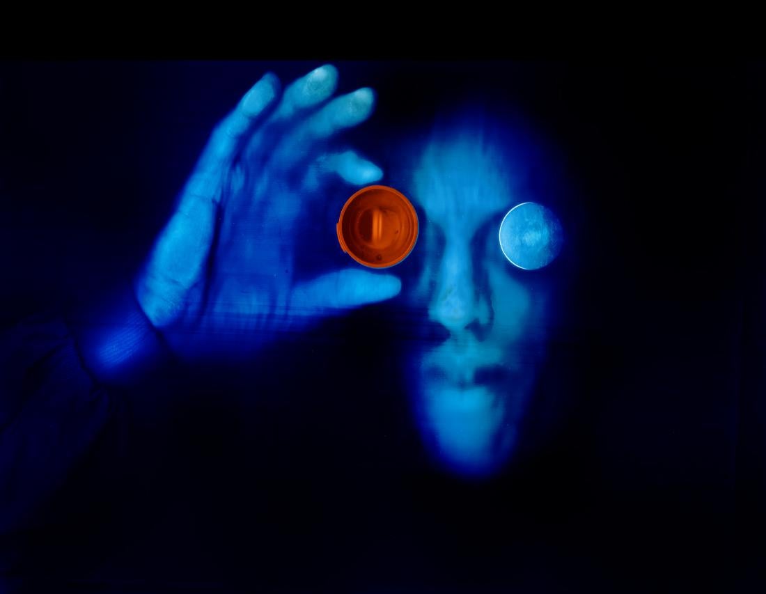 A color self- portrait scanograph. I am featured in blue with a black background. My right eye is covered with a glass lens also colored in blue. My right hand is lifted holding an orange funnel over my right eye. This image is from my “Transmog Project” body of work.