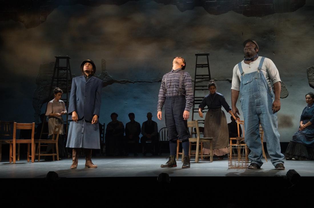 A black female cop, a white male with dark pants and plaid shirt with suspenders, and a black male in overalls are looking up.