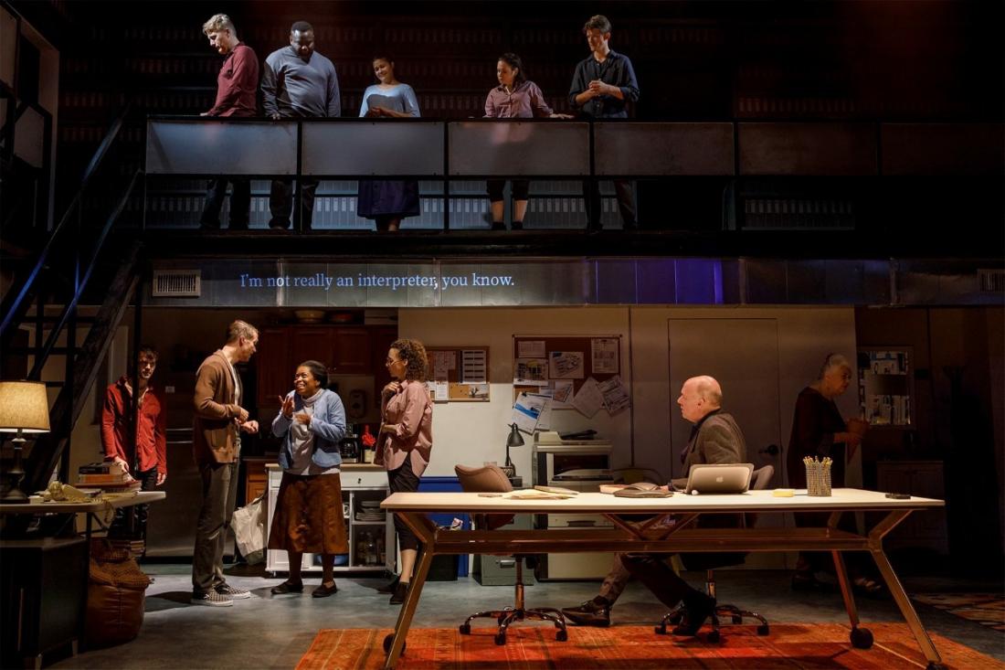 On the stage, there are six people starting with a whilte male in red shirt, a white male with brown cardigan looking at a black female as they talked along with a white female with curly hair looking at them,  An older male sitting by a table with a laptop in front of him looking at them and an older woman standing in the corner. 