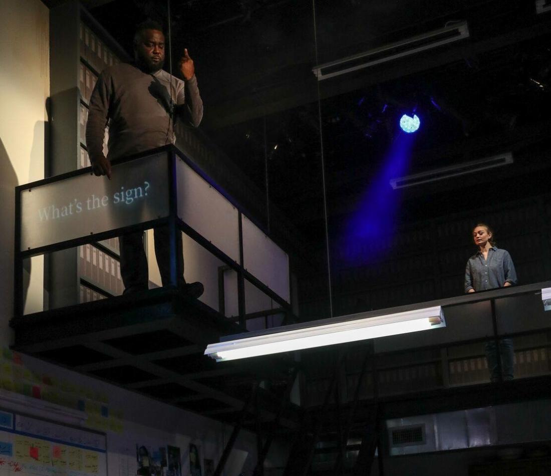 A black male standing and looking down at the stage along with a white female standing nearby. The sign in front of the black male: "What's the sign?"