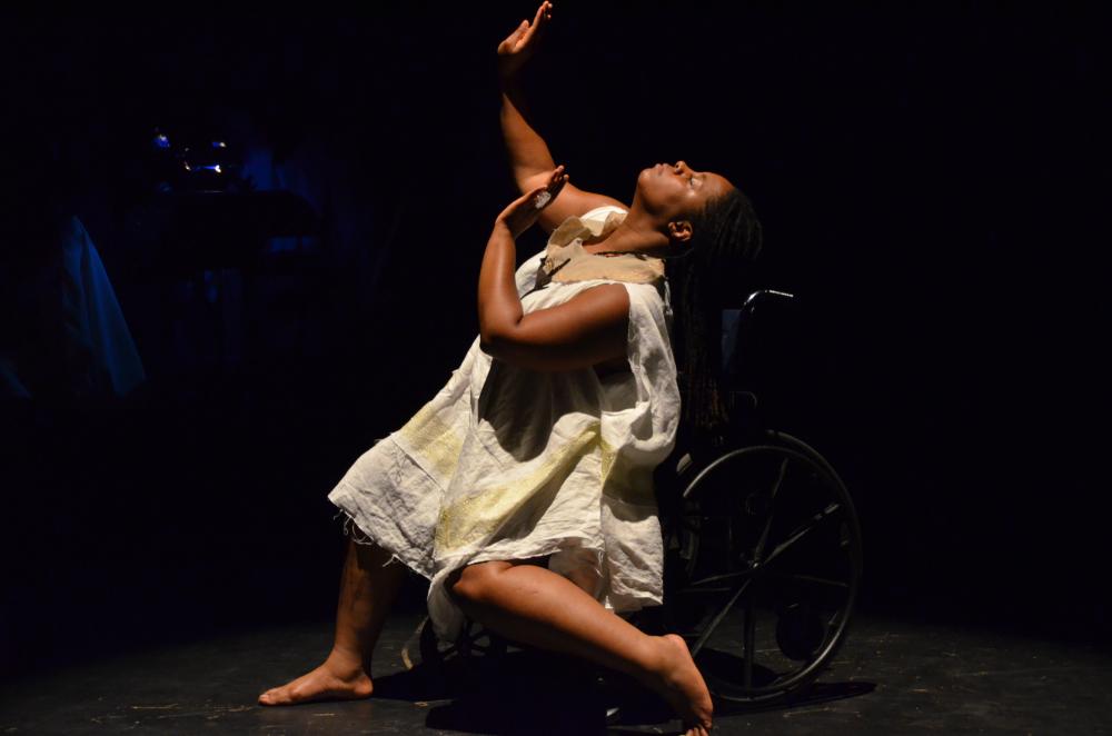 India Harville, African American woman barefoot with black locs wearing a loose white dress. Against a dark background, she is dancing in her manual wheelchair posing sideways with hands reaching towards the sky and left knee bent.