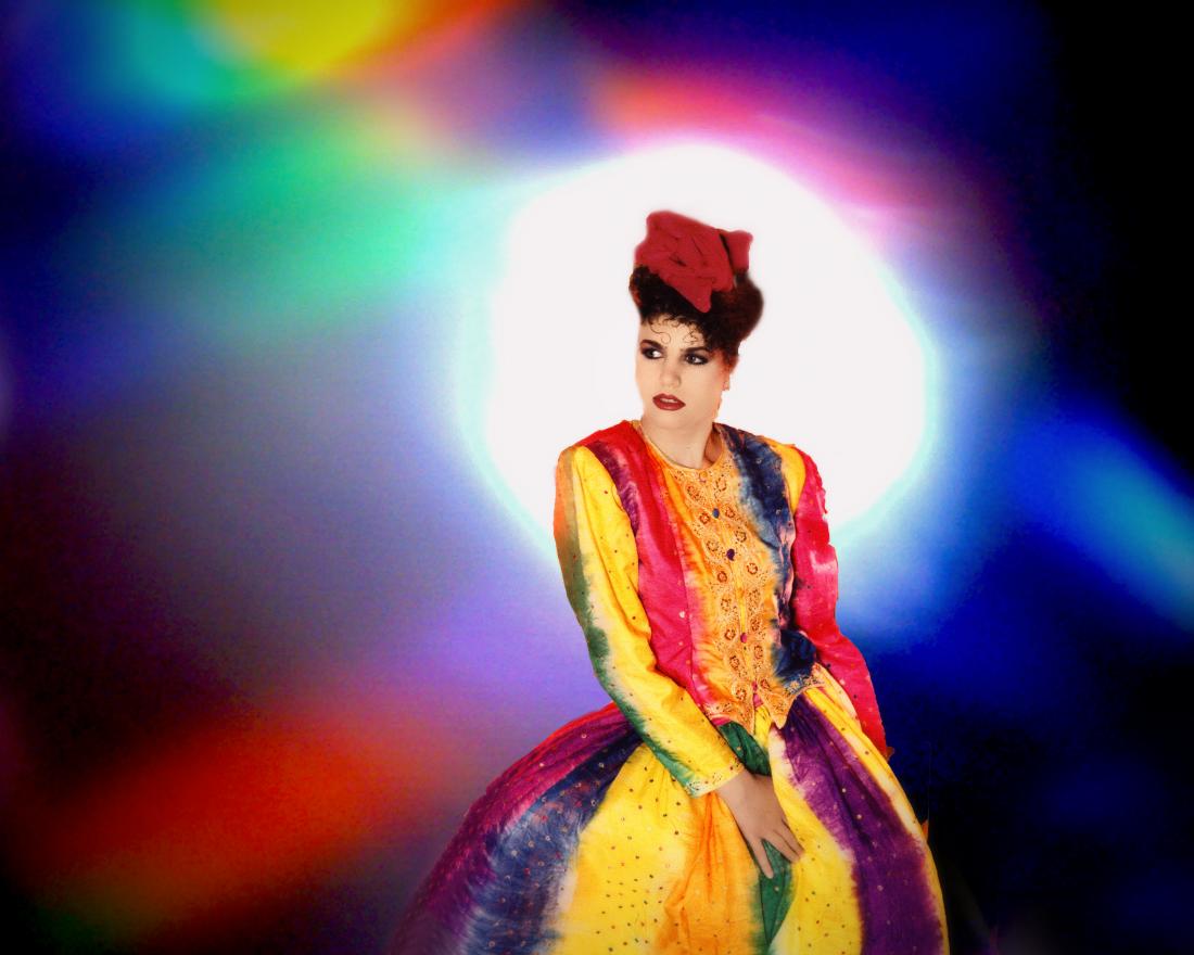A photomontage of a runway model wearing a rainbow-colored dress. Her torso is twisting as she pivots the runway. A spotlight beam positioned directly behind her head enshrouds her in a halo. The light splinters and fragments into various rainbow colors. This image is from my “Neo Glam” series of reimagined fashion portraits.