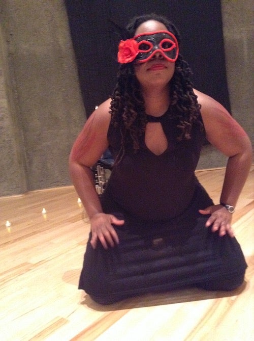 India Harville, African American woman barefoot  with long black curly locs wearing a black dress with a red/black face mask and a red flower on the right side. She is kneeling on a brown wooden floor surrounded by white electric candles and a gray/black wall. She is dancing with her hands on her thighs.