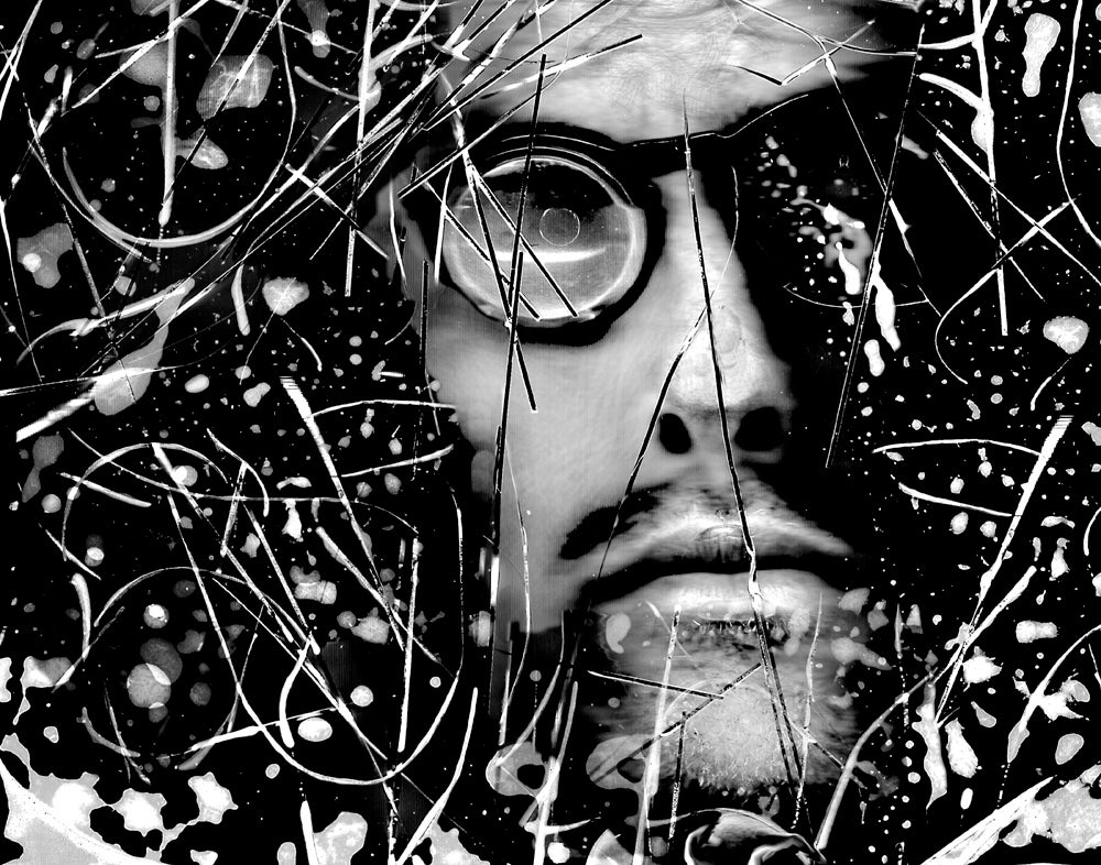 A black & White self-portrait scanograph. My face is looking directly at the viewer. I am wearing thick black framed magnifying glasses. There is debris and detritus floating over my face and throughout the frame. These obstructions represent the floaters I experience in my own vision. This image is from my “Blind Vision” series. 