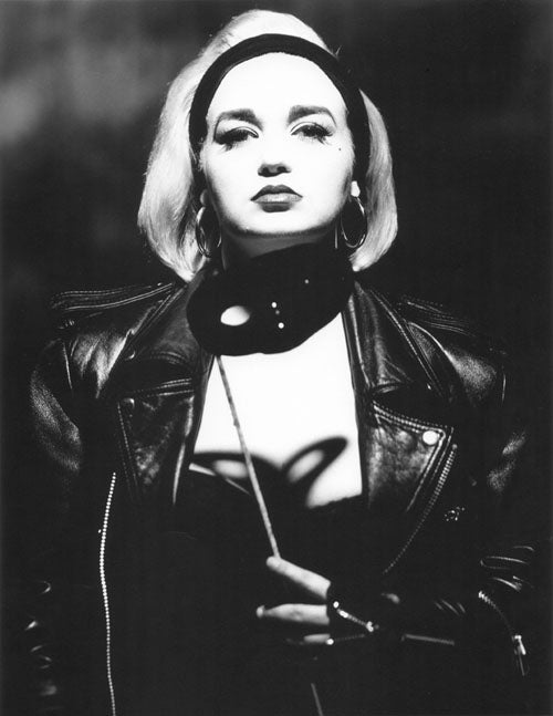 A Black & White photographic portrait of a female model.  She is wearing a black hair band pushing back her platinum blond hair. She is wearing a black leather jacket which is open revealing some of her torso. She is holding a mask near her chin. It is attached to a stick. The studio light from above is casting a shadow of the mask onto her torso. The shadow looks like a butterfly.