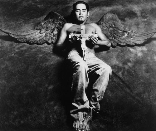 A photographic portrait of a young Latino. He is laying on a fabric background. He is shirtless and holding a crucifix over his torso. Behind him are angel’s wings which are the same color as the background fabric. His eyes are closed and a beautiful white light is illuminating his wings and body. This image is from my “Remember – an AIDS memorial retrospective.” Body of work.