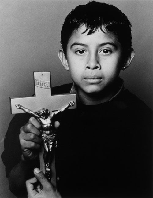 A Black & White photographic portrait of a young Latin boy holding a crucifix. He is staring directly into the camera with beautiful dark eyes. He has a somber expression. Both of his parents have died from AIDS.  This portrait is from my “Remember – an AIDS memorial Retrospective” body of work. 