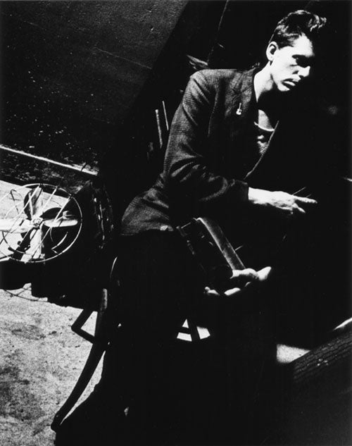 A Black & White photographic portrait of a young man seated on a chair in front of a black wall. He is wearing a black sport coat and holding piano pedals in his hands. He is looking off camera to the right side of the frame. He has a small pin in the shape of a guitar affixed to his lapel The image is photographed on the diagonal creating tension within the frame. On the white concrete floor to his left is a vintage metal circular fan. This image is from my “Remember – an AIDS Memorial Retrospective” body 