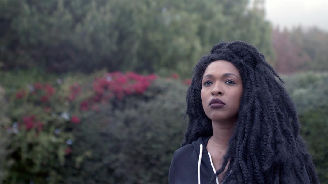 A Black woman with dreds, wearing a black zip up sweatshirt, looks over and past the camera. She stands in front of green bushes with pink flowers shrouded in fog.