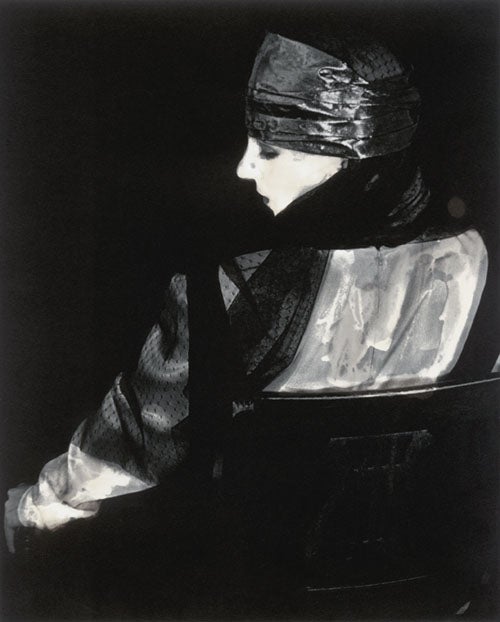 A Black & White photographic portrait of a young woman seated on a chair. The woman and chair is turned with their backs toward the camera.  The woman has her face turned so you can see her profile. She is wearing a silvery fabric gown. The same fabric is wrapped around her head as a hair piece. She has black batiste fabric draped over her shoulders and running down her left arm. The style of the gown and pose resembles an image that would have appeared in a 1920’s Vogue magazine.
