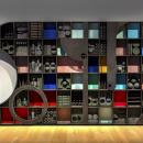 A large sculpture made of many rectangular cubbies filled with shaped acoustic foam stands flat against a wall. The depth of the cubbies casts shadows over flat red, blue, yellow, aqua, and pink foam panels. Flat, black pieces of wood in various shapes cover parts of the sculpture. The left side has a half circle cut out with a musical triangle hanging inside.