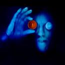 A color self- portrait scanograph. I am featured in blue with a black background. My right eye is covered with a glass lens also colored in blue. My right hand is lifted holding an orange funnel over my right eye. This image is from my “Transmog Project” body of work.