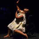 India Harville, African American woman barefoot with black locs wearing a loose white dress. Against a dark background, she is dancing in her manual wheelchair posing sideways with hands reaching towards the sky and left knee bent.