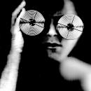 A Black & White scanographic self-portrait from my “Blind Vision” series.  My face appears blurred and distorted. I am holding film reels over my eyes. The film reels represent the magnifiers I use on a constant basis in order to negotiate my life as a legally blind artist.
