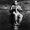 A photographic portrait of a young Latino. He is laying on a fabric background. He is shirtless and holding a crucifix over his torso. Behind him are angel’s wings which are the same color as the background fabric. His eyes are closed and a beautiful white light is illuminating his wings and body. This image is from my “Remember – an AIDS memorial retrospective.” Body of work.