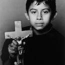 A Black & White photographic portrait of a young Latin boy holding a crucifix. He is staring directly into the camera with beautiful dark eyes. He has a somber expression. Both of his parents have died from AIDS.  This portrait is from my “Remember – an AIDS memorial Retrospective” body of work. 