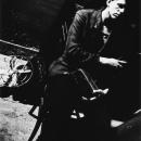 A Black & White photographic portrait of a young man seated on a chair in front of a black wall. He is wearing a black sport coat and holding piano pedals in his hands. He is looking off camera to the right side of the frame. He has a small pin in the shape of a guitar affixed to his lapel The image is photographed on the diagonal creating tension within the frame. On the white concrete floor to his left is a vintage metal circular fan. This image is from my “Remember – an AIDS Memorial Retrospective” body 