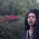 A Black woman with dreds, wearing a black zip up sweatshirt, looks over and past the camera. She stands in front of green bushes with pink flowers shrouded in fog.