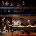 On the stage, there are six people starting with a whilte male in red shirt, a white male with brown cardigan looking at a black female as they talked along with a white female with curly hair looking at them,  An older male sitting by a table with a laptop in front of him looking at them and an older woman standing in the corner. 