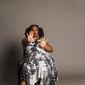 India Harville, African American woman barefoot with black locs in two ponytails and face glitter dancing against a gray background. She is wearing a long black/white dress dancing in her manual wheelchair with both face and palms facing forward with all fingers spread out.